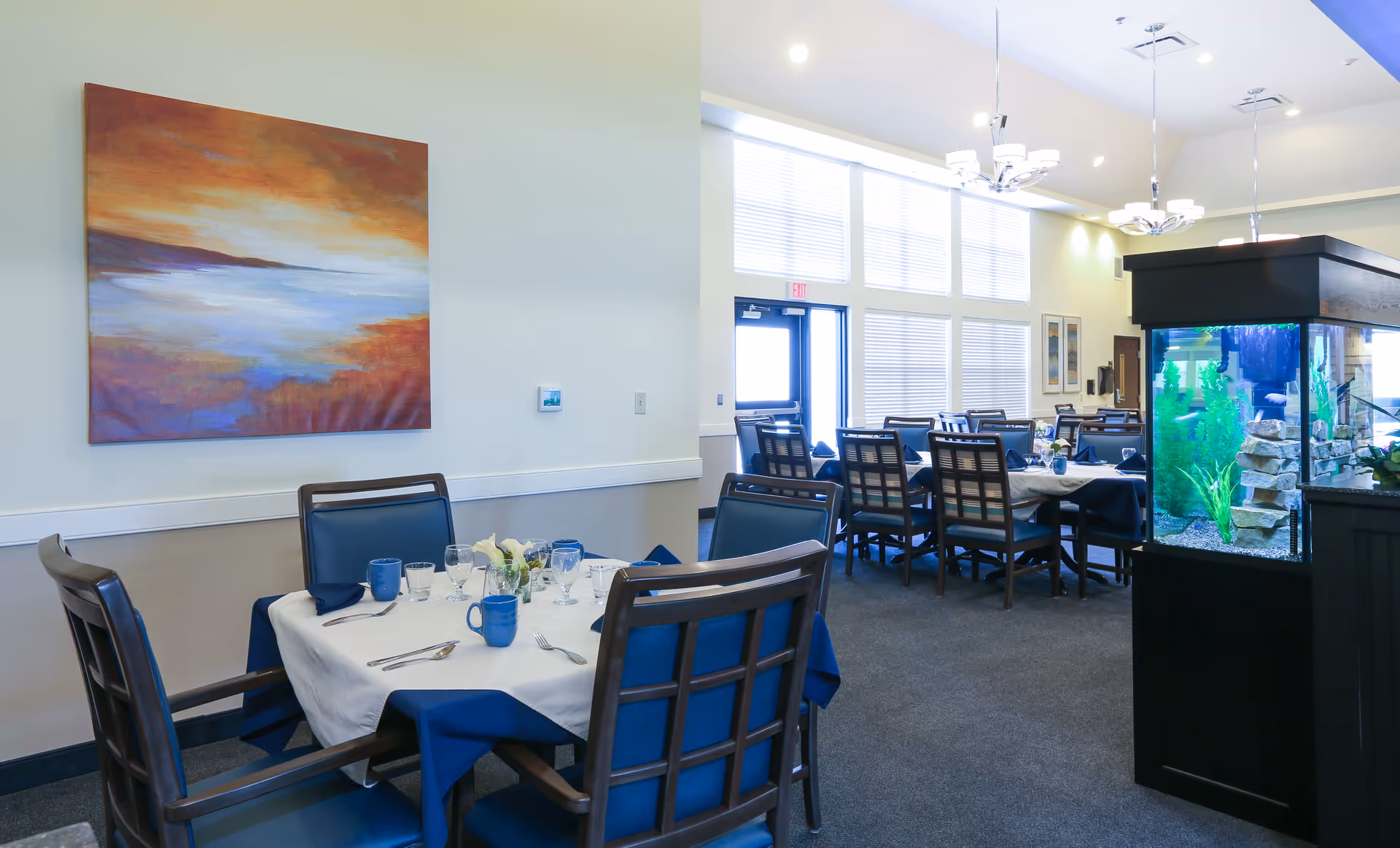 A bright dining room with tables covered in white and blue tablecloths, set with blue mugs, glassware, and silverware. There are wooden chairs with blue cushions around the tables. A large colorful painting hangs on the wall, and a large fish tank with green plants and rocks is visible on the right side. Large windows with blinds allow natural light to fill the room.