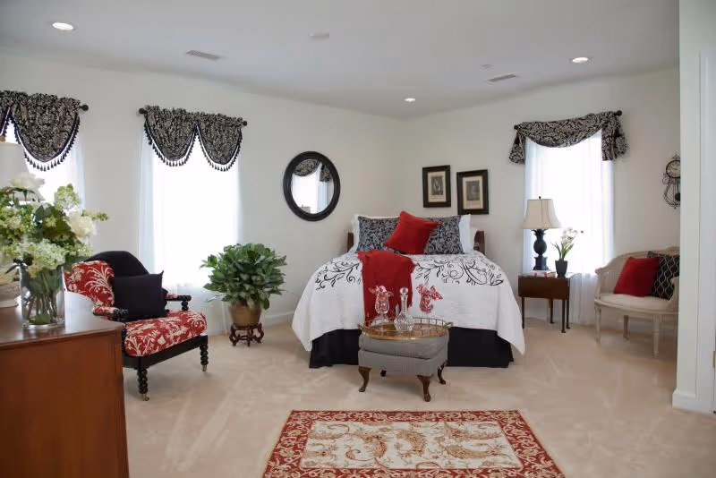 A well-decorated bedroom with a bed centered against the wall, adorned with white and black bedding and red accent pillows. There is a small ottoman with a tray holding glassware at the foot of the bed. The room features two windows with patterned valances, a round mirror on the wall, framed pictures, a wooden dresser with a vase of flowers, a red and black patterned armchair, a green potted plant, a side table with a lamp and flowers, and a small bench with red and black pillows. The floor is carpeted with a decorative area rug in front of the bed.
