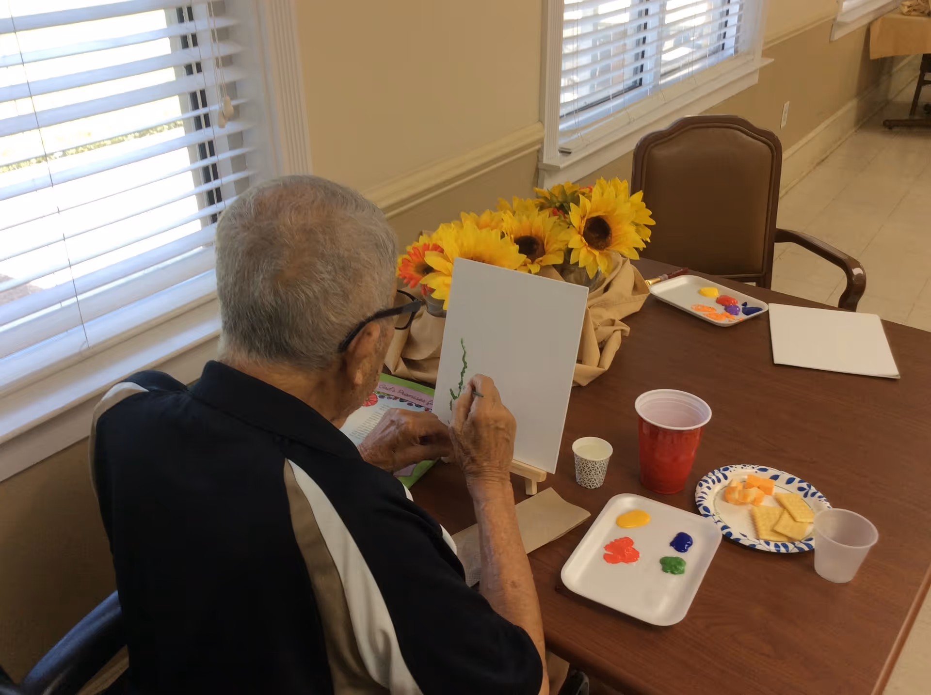 An elderly man with gray hair and glasses is seated at a table by a window with blinds, painting on a small canvas. On the table are paint palettes with various colors, a red plastic cup, a paper plate with crackers and cheese, and a bouquet of sunflowers in the background.