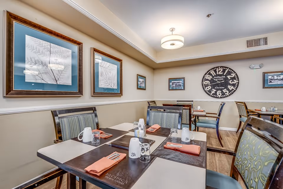 Dining room in a senior living facility with several tables set with placemats, napkins, cups, and glasses. The walls are decorated with framed artwork and a large vintage-style clock. The room has a warm and inviting atmosphere with soft lighting and wooden flooring.