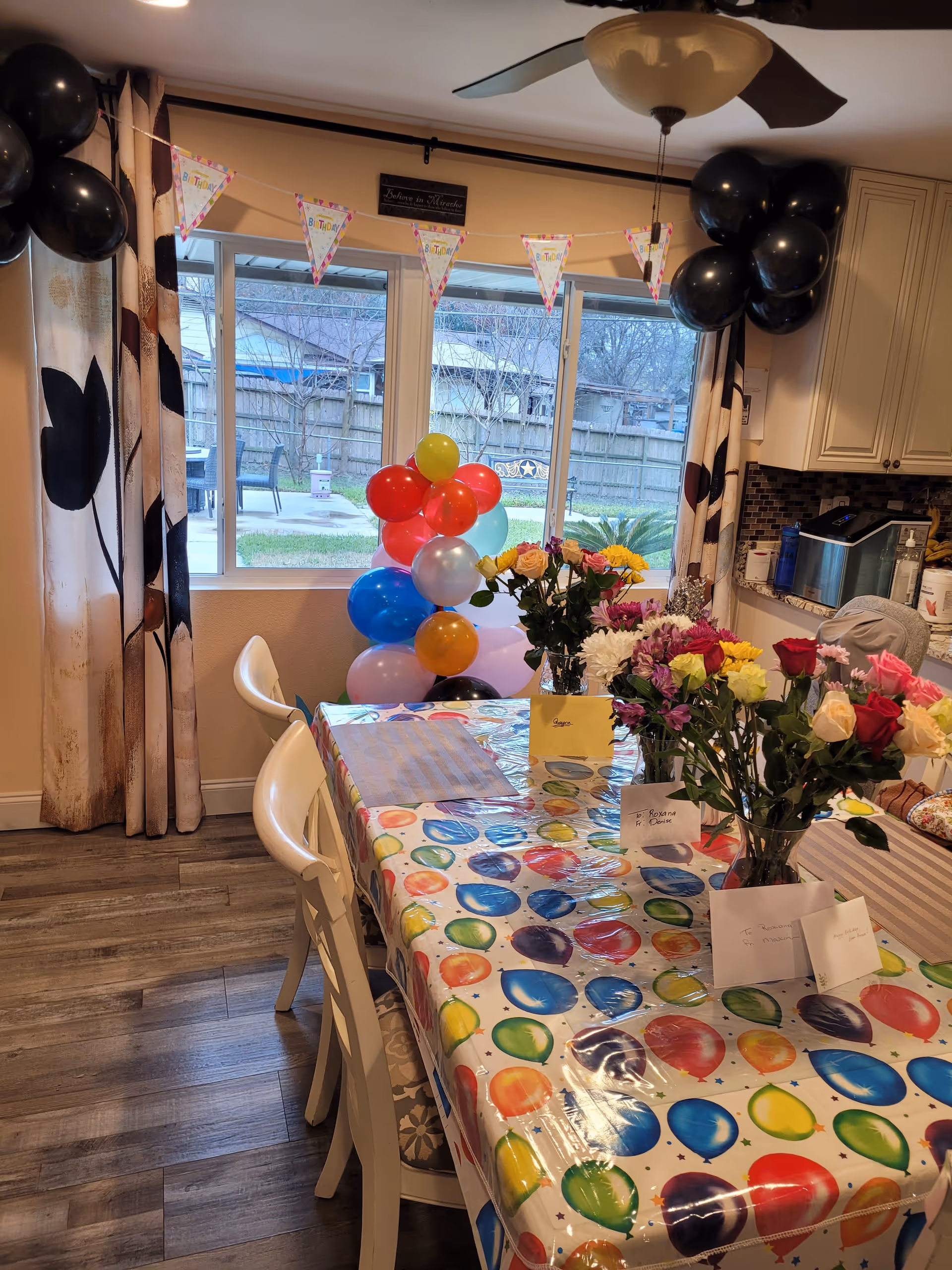 A decorated dining area with a table covered in a colorful balloon-patterned tablecloth. The table has several flower bouquets in vases and greeting cards. There are black balloons attached to the curtain rod and a string of triangular birthday banners hanging across the window. Outside the window, a fenced backyard with patio furniture is visible. The room has wooden flooring and light-colored walls with patterned curtains.