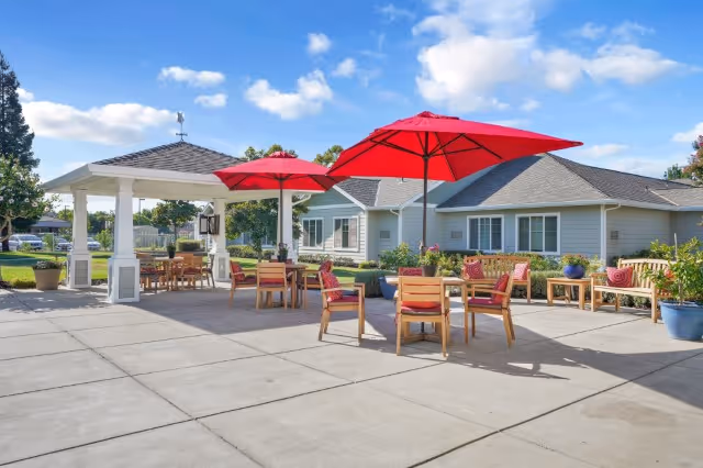 Outdoor patio area at The Commons on Thornton with wooden tables and chairs, red umbrellas providing shade, a gazebo, and a building with light gray siding in the background under a partly cloudy blue sky.