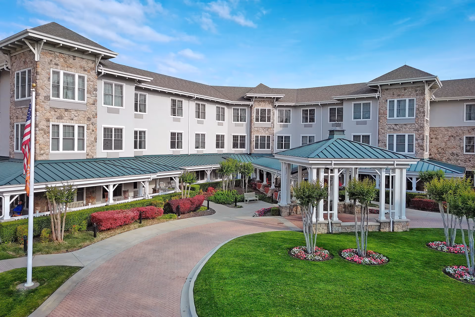 Exterior view of a senior living facility with a three-story building featuring stone and light-colored walls, numerous windows, a covered walkway with a green metal roof, a gazebo with a matching roof, well-maintained landscaping including green grass, flower beds, and small trees, and an American flag on a flagpole.