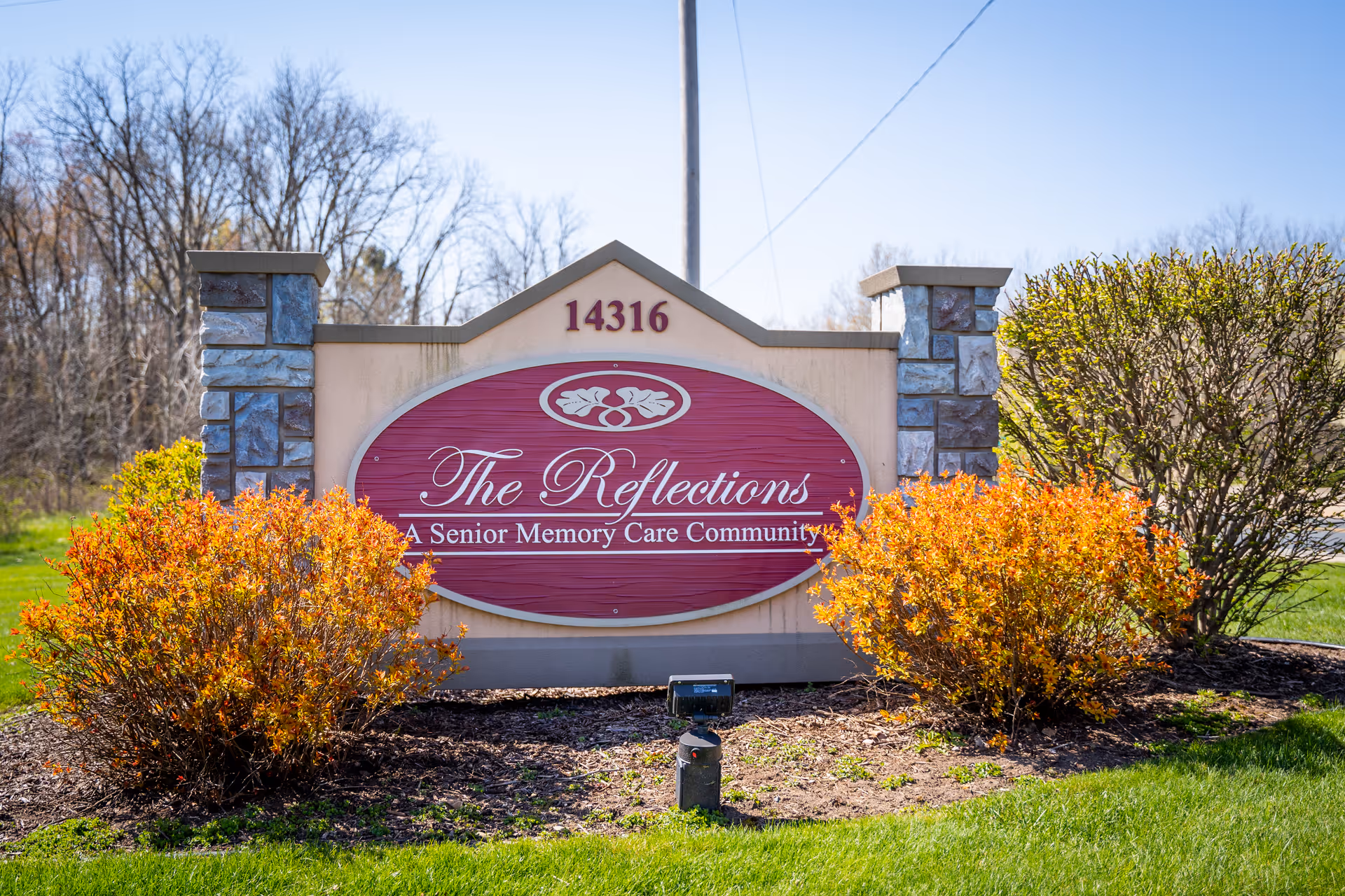 Entrance sign reading "The Reflections A Senior Memory Care Community" with address 14316 set between stone pillars and landscaped bushes.