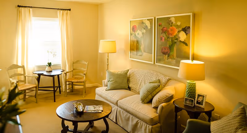 A cozy living room with a beige sofa adorned with green pillows and a knitted throw. Two framed floral paintings hang above the sofa. There is a round wooden coffee table with a decorative item and a magazine on it. To the right of the sofa is a side table with a green lamp and framed photos. Two wooden chairs with woven seats and a small round table are placed near a window with white curtains. The room is warmly lit by natural light and lamps.