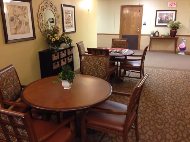 Interior view of a senior living facility common area with two round wooden tables surrounded by patterned cushioned chairs. One table has a small potted plant, and the other has a checkers game set up. The walls are decorated with framed artwork and a decorative mirror above a cabinet with baskets. In the background, there is a wooden door, a console table with decorative items, and an exit sign above the door.