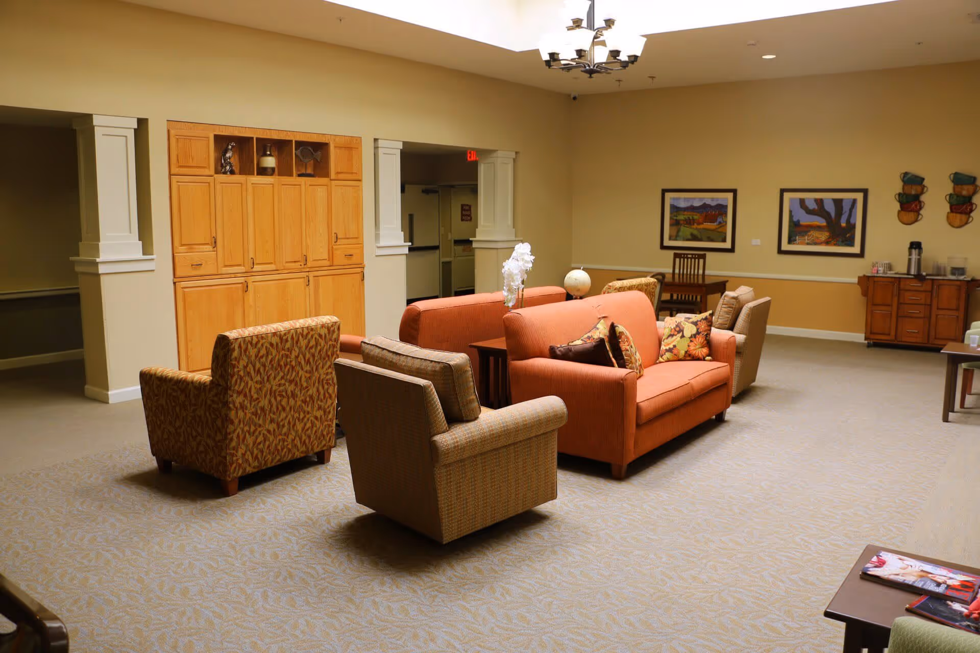 A spacious living room area in a senior living facility with beige walls and carpeted floor. The room features two orange sofas with floral cushions, two patterned armchairs, a wooden cabinet, a small table with a globe and a white orchid, framed artwork on the walls, and a sideboard with a coffee station. The ceiling has a modern chandelier and a skylight.