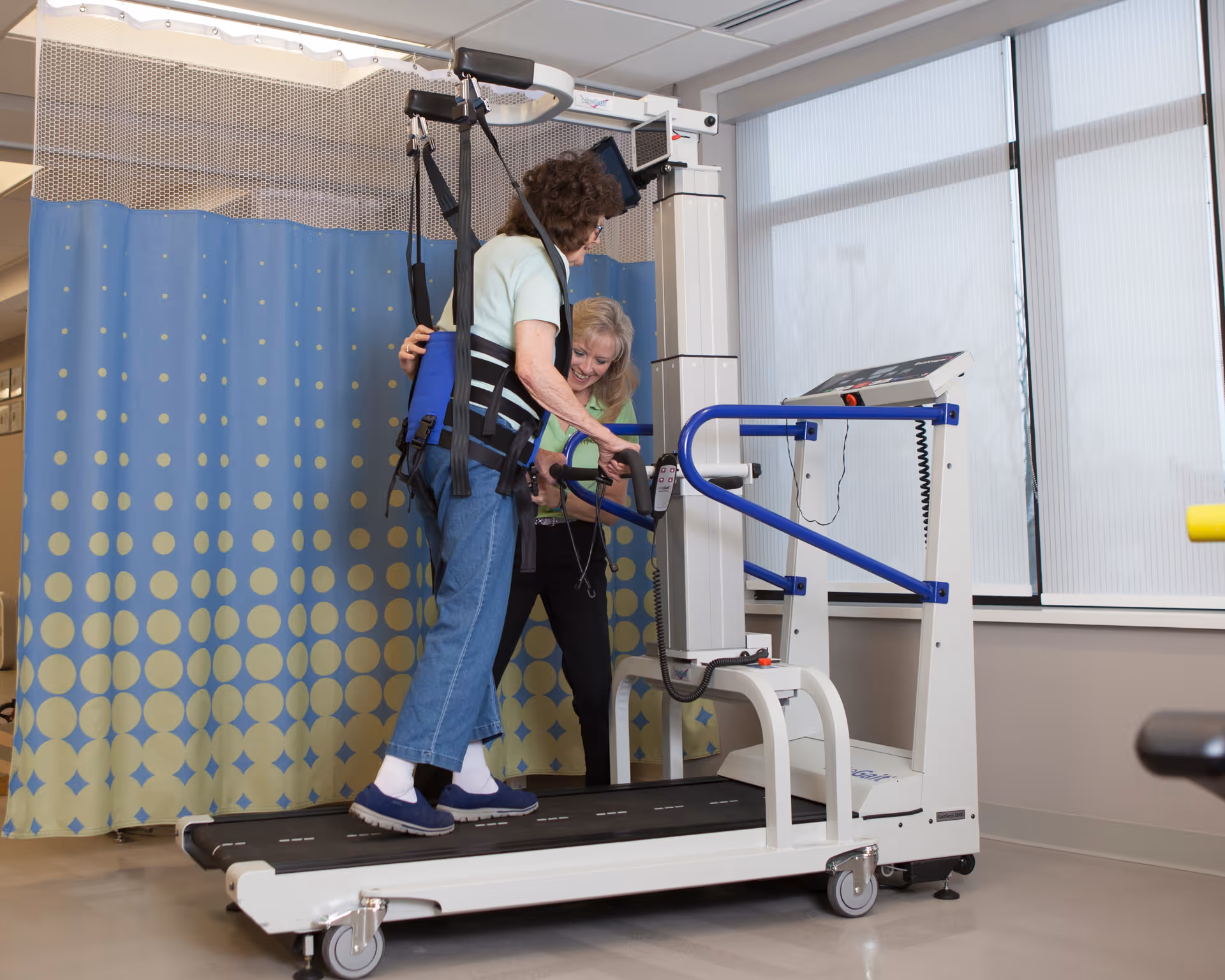 An elderly woman wearing a support harness is assisted by a caregiver while walking on a treadmill in a rehabilitation or physical therapy room with a blue and yellow patterned privacy curtain and large windows.