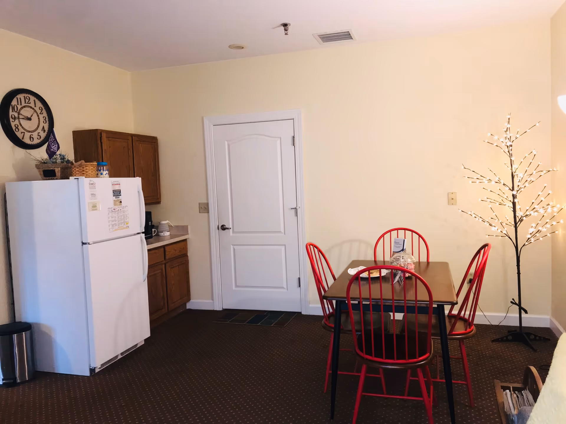 Small dining area with a wooden table and red chairs next to a kitchenette with a white refrigerator and a decorative lit tree.