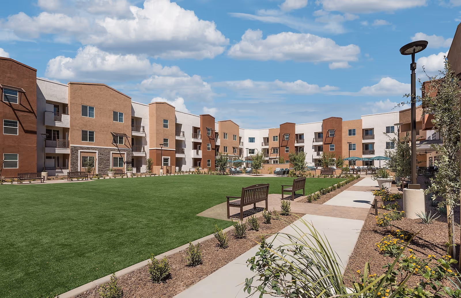Outdoor courtyard area of a senior living facility with a large green lawn, benches, landscaped plants, and a multi-story building with balconies surrounding the space under a partly cloudy sky.