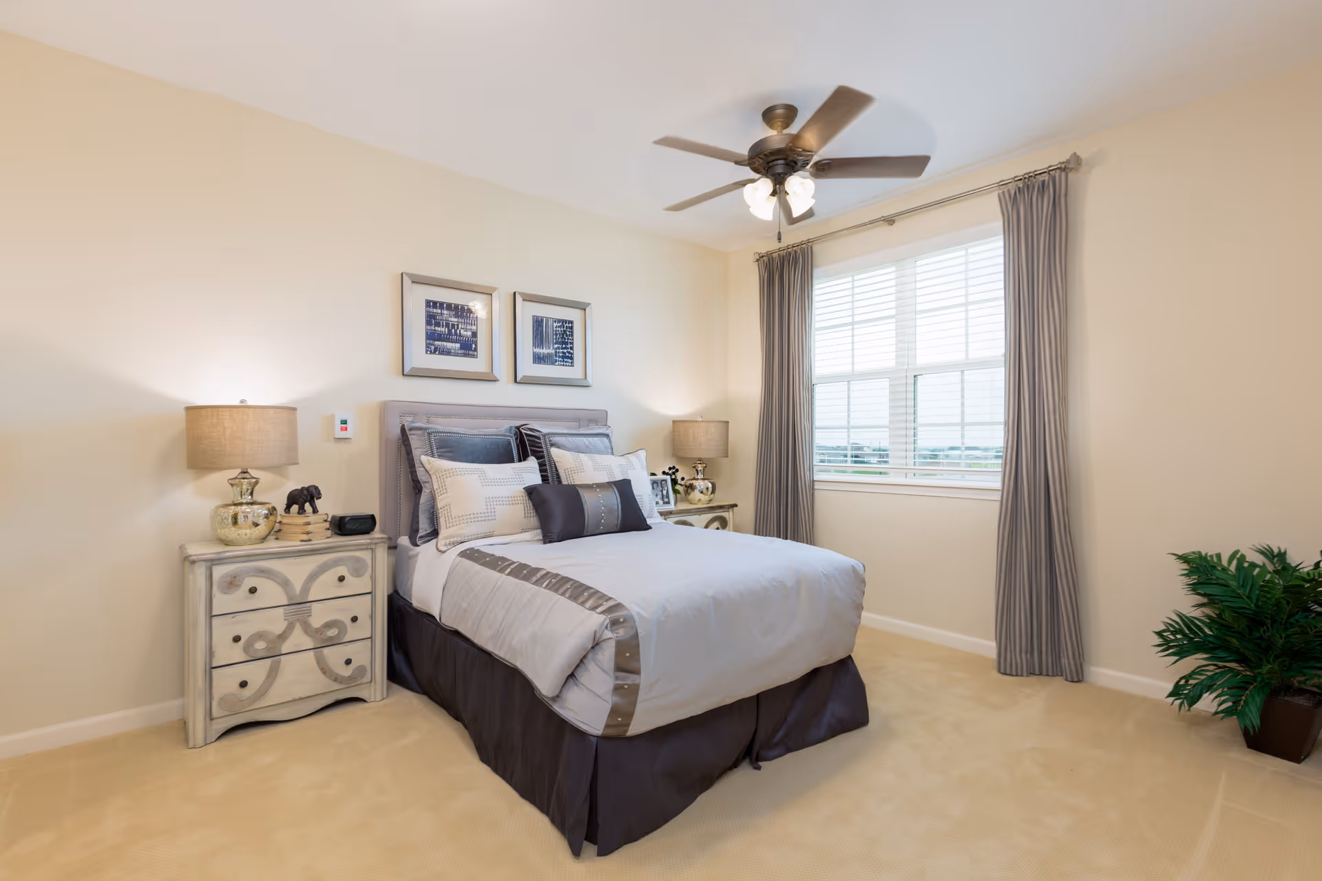 A neatly made bed with multiple pillows in a bright bedroom featuring beige walls, a ceiling fan with lights, two bedside tables with lamps and decorative items, a large window with gray curtains, and a potted plant in the corner.