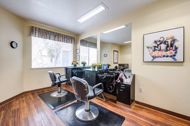 Interior of a salon area in a senior living facility with two black salon chairs facing a large mirror. There are two black hair washing sinks with cabinets underneath, hair care products on the counter, and a colorful painting of two elephants in a bathtub on the wall. The room has wood flooring, a window with patterned valance, and a wall clock.