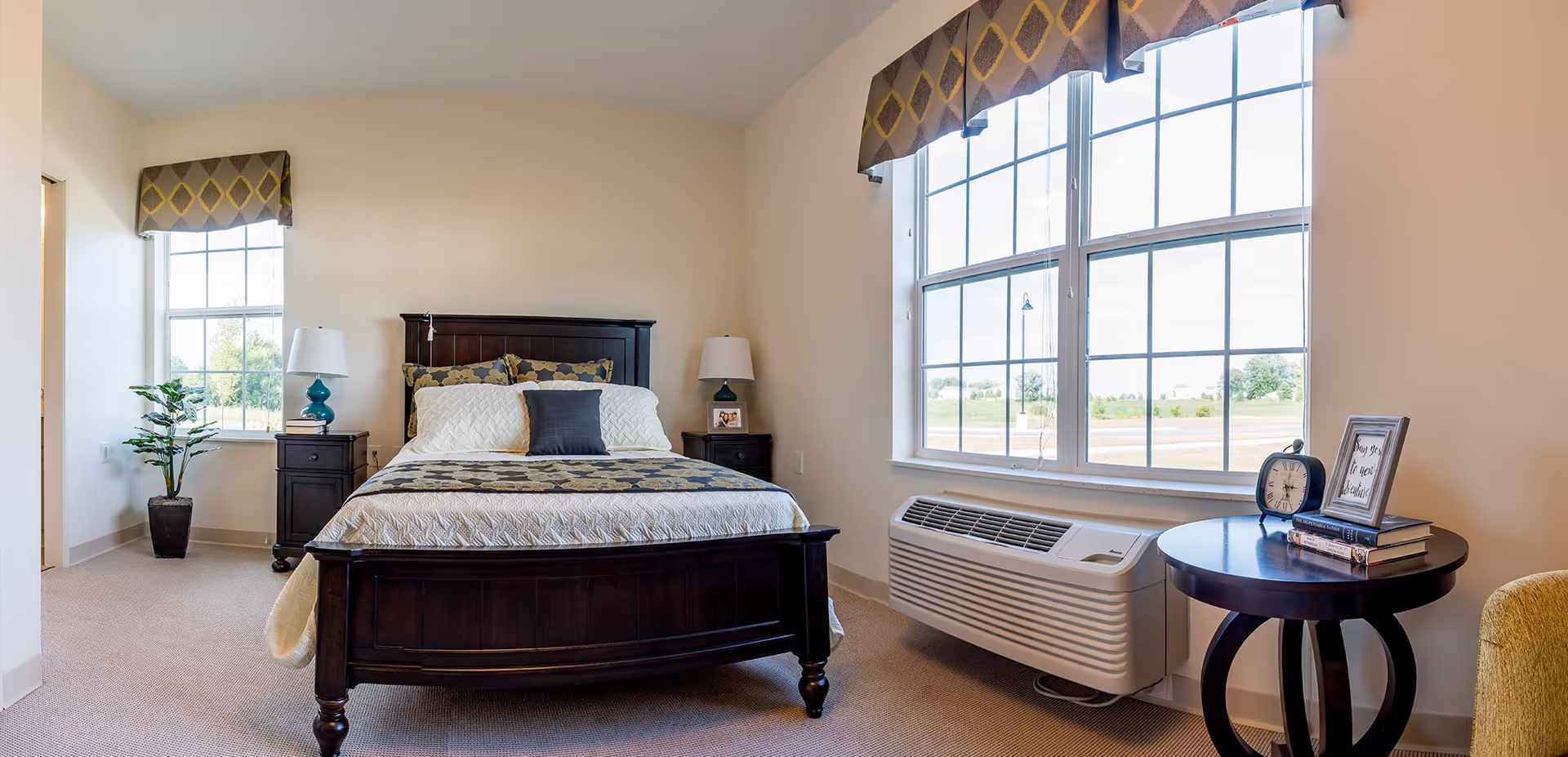 A bright bedroom with a dark wooden bed frame and a neatly made bed with patterned pillows and a quilt. There are two windows with patterned valances, a potted plant near one window, two bedside tables with lamps, and a round side table with a clock, books, and a framed quote. An air conditioning unit is installed under one window.