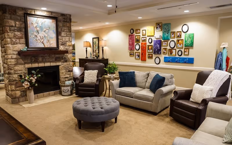 A cozy living room with a stone fireplace on the left wall, a large painting above the fireplace, and a variety of seating including a beige sofa with blue pillows, two brown armchairs with white pillows, and a gray tufted ottoman in the center. The walls are decorated with colorful framed artwork and photos, and there are lamps and plants adding to the warm ambiance.