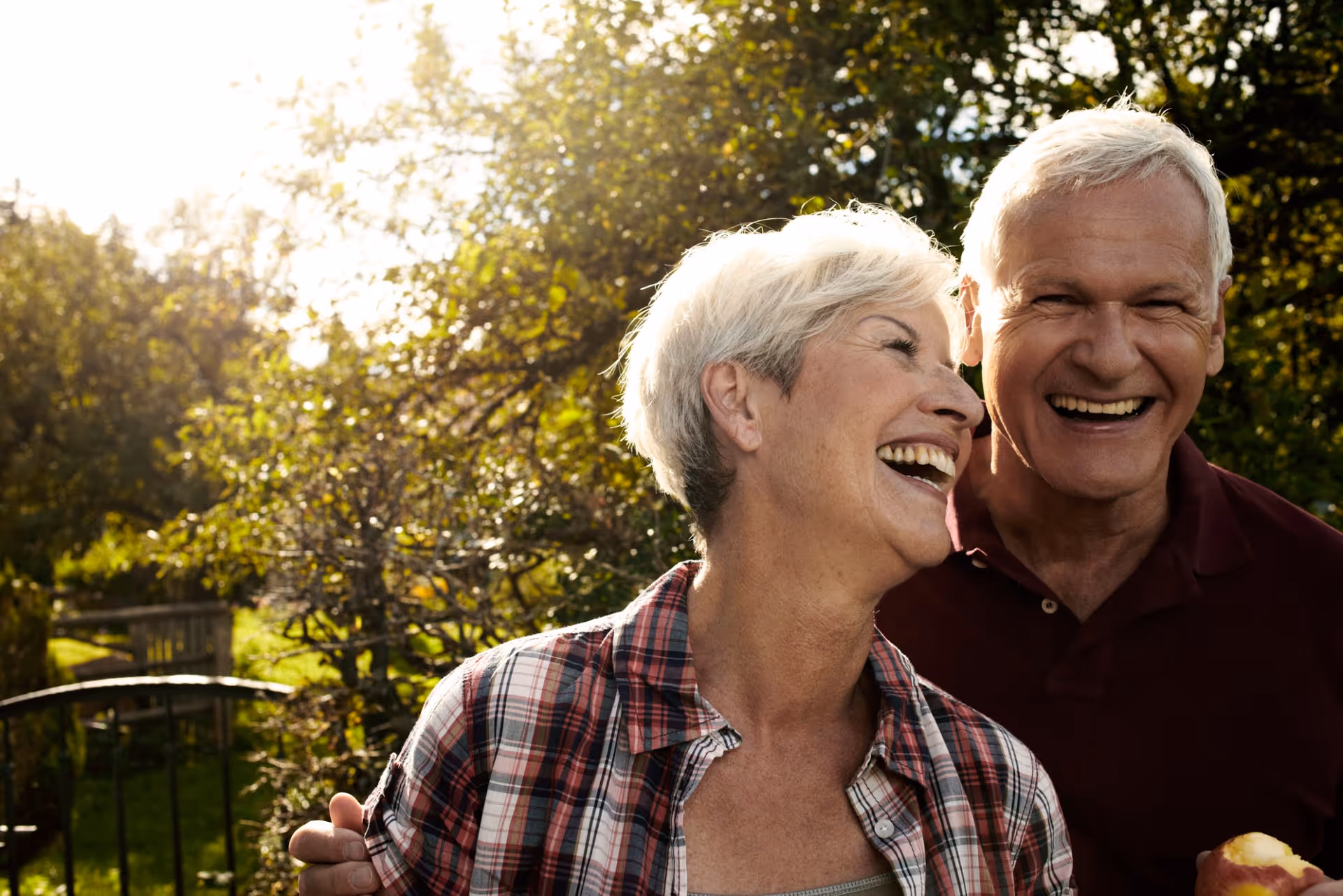 An elderly couple outdoors in a garden or park area, smiling and laughing together with sunlight filtering through the trees in the background.