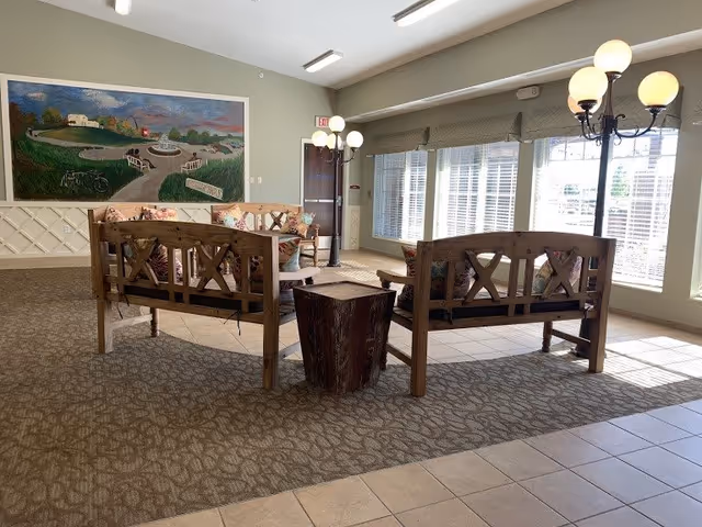 A cozy seating area in a senior living facility with wooden benches featuring floral cushions arranged around a small wooden stump table. The room has large windows with blinds allowing natural light to fill the space, a patterned carpet, and a mural painting on the wall depicting a scenic outdoor landscape. There are also tall floor lamps with round white globes providing additional lighting.