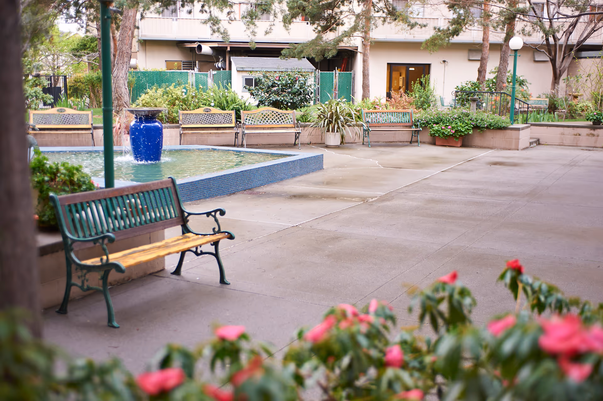 Outdoor courtyard with benches, a blue fountain, planters, and flowering shrubs in front of a building.