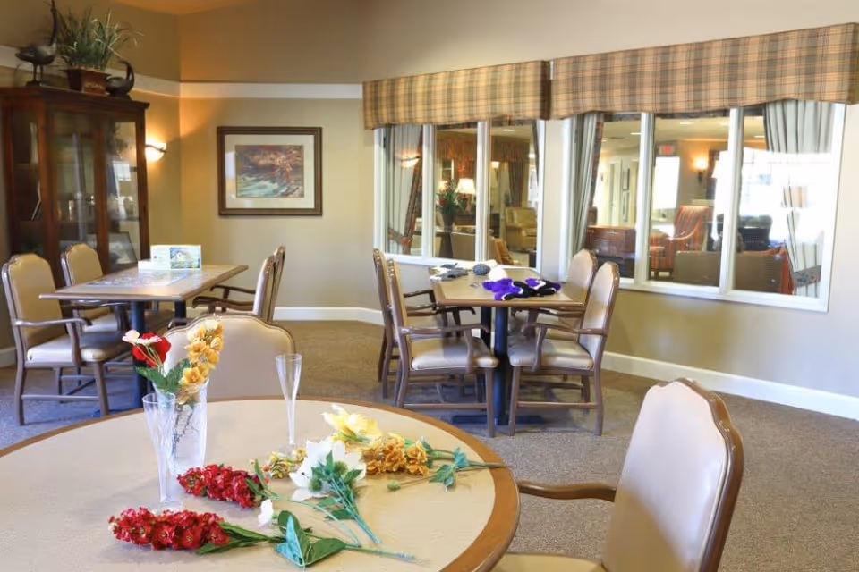 A cozy dining area with several tables and chairs arranged around the room. One table in the foreground has artificial flowers and two empty glasses. The walls are painted beige with a framed picture hanging, and a large window with plaid valances looks into an adjacent room with more seating. A wooden cabinet with glass doors is visible in the corner.