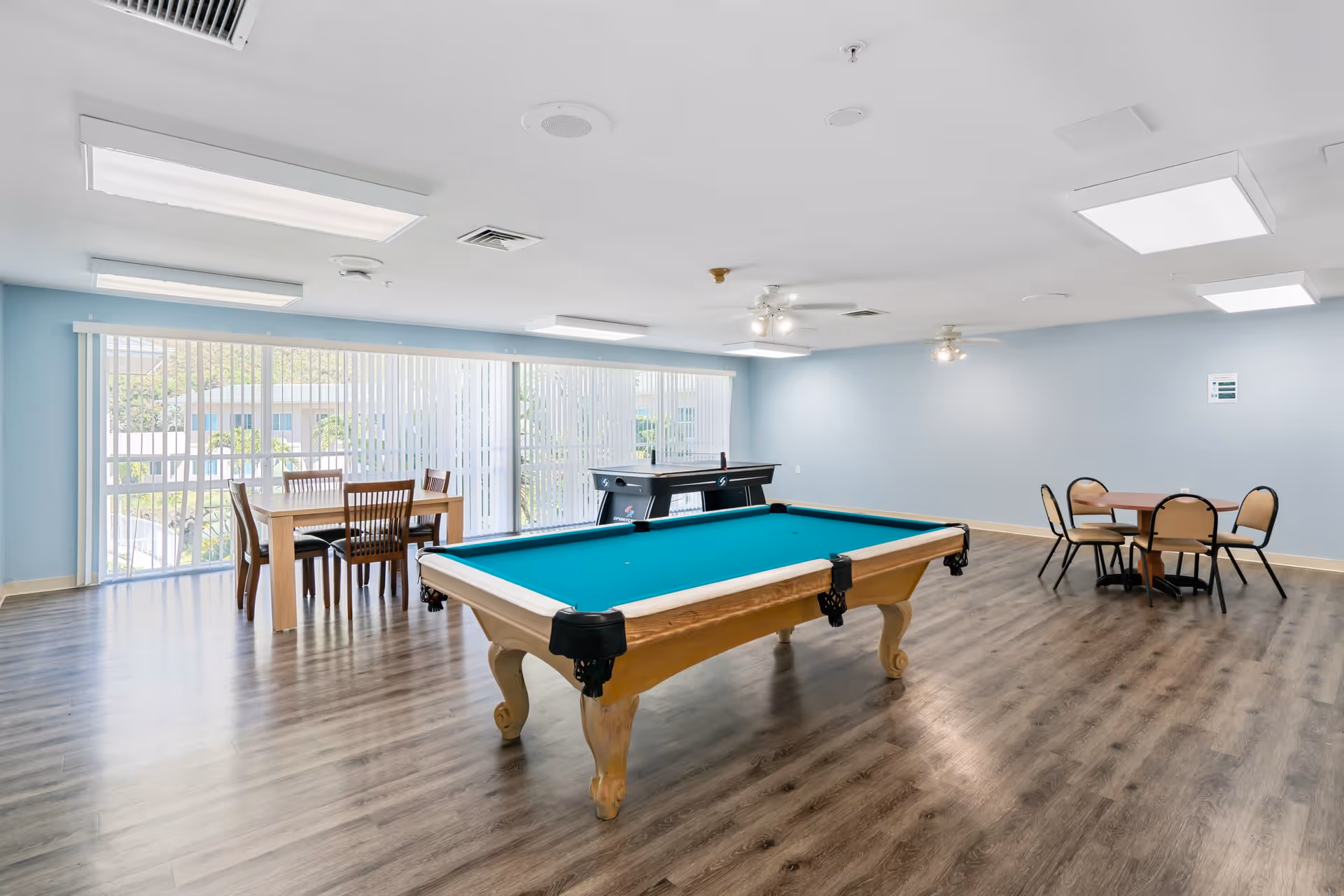 A spacious recreational room with a pool table in the center, an air hockey table near large windows with vertical blinds, a wooden dining table with four chairs on the left, and a round table with four chairs on the right. The room has light blue walls, wood flooring, and ceiling lights.