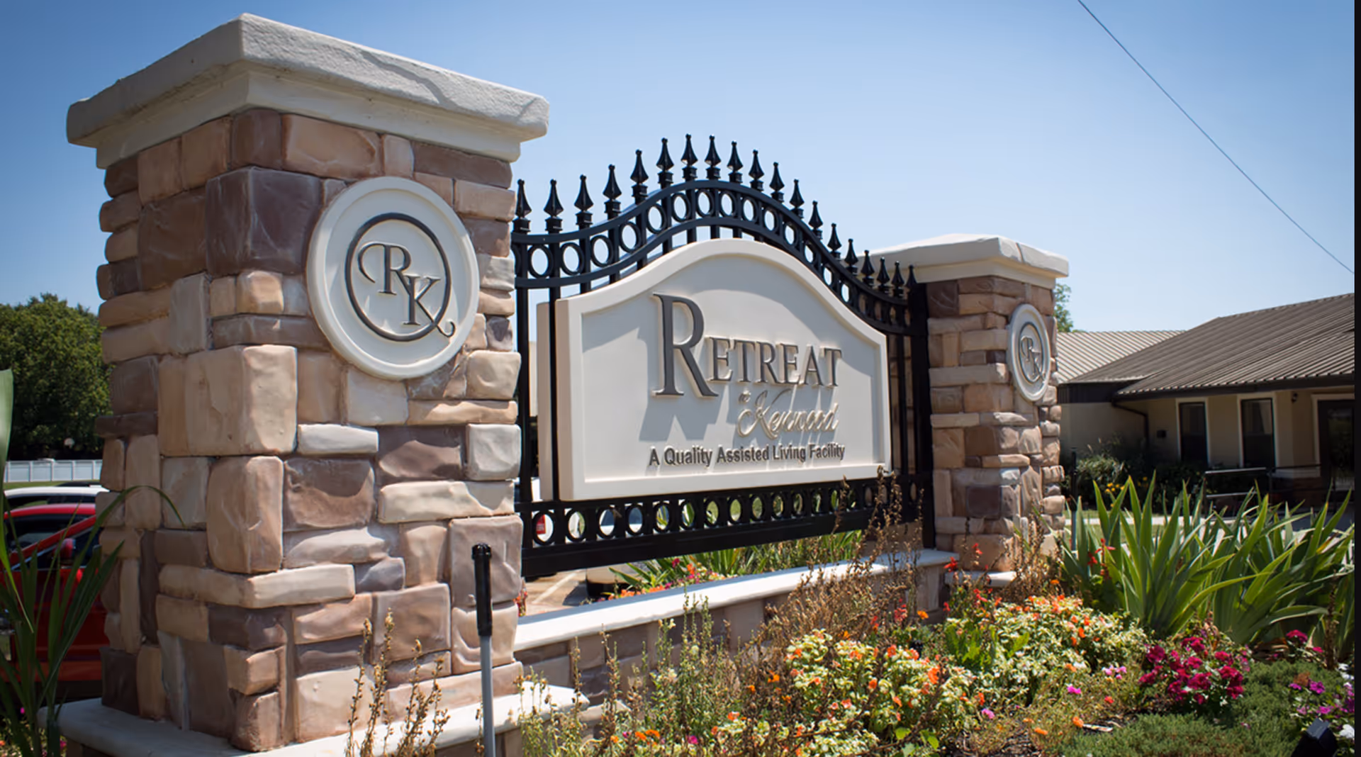 Stone and wrought-iron entrance sign for The Retreat at Kenwood surrounded by flowers with the facility building in the background.