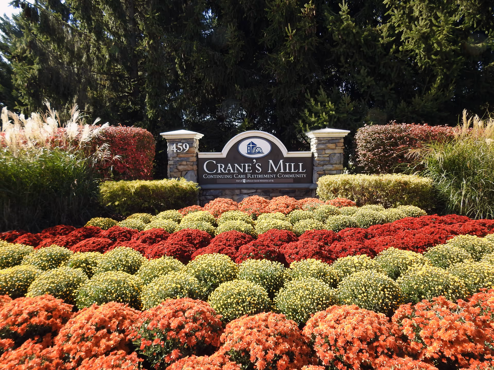 A landscaped garden area with rows of colorful flowers in red, yellow, and orange in front of a stone sign that reads 'Crane's Mill Continuing Care Retirement Community' with the address number 459 on a stone pillar to the left.