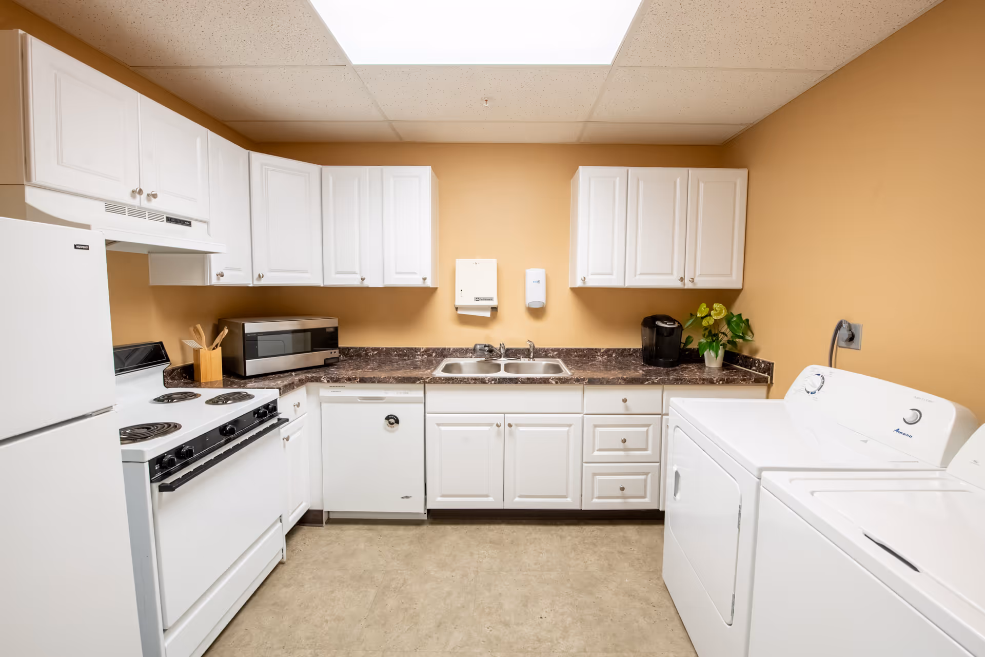A clean kitchen area with white cabinets, a white refrigerator, stove, microwave, dishwasher, and a double sink. There is a coffee maker and a small potted plant on the countertop. To the right, there is a white washing machine and dryer. The walls are painted beige and the floor is a light-colored linoleum.