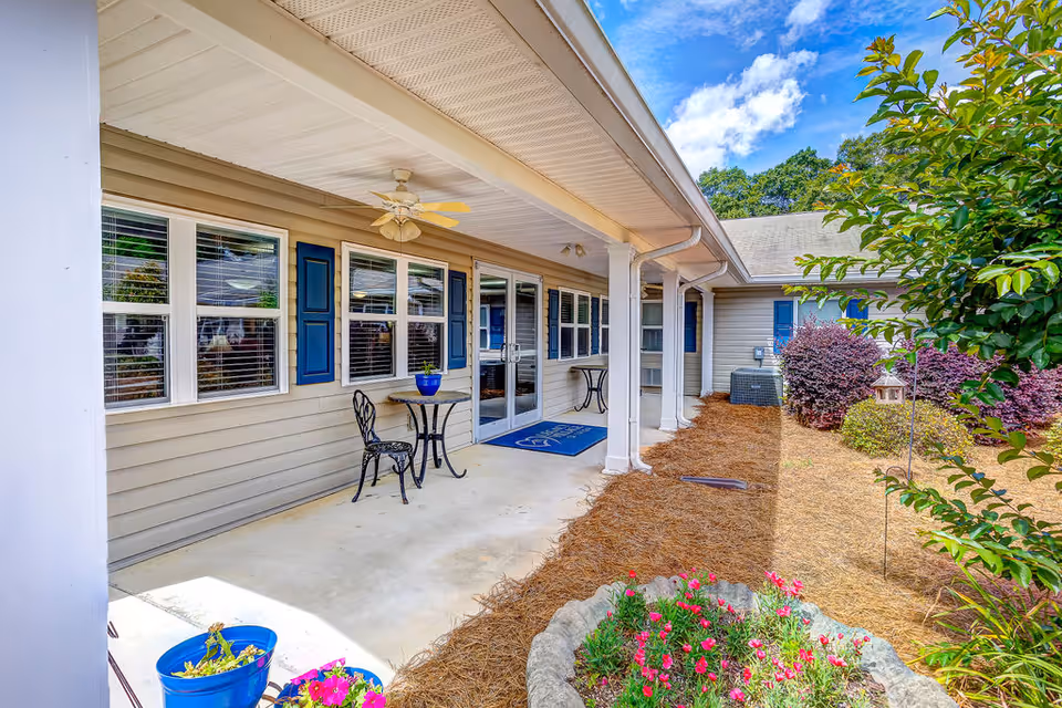 Outdoor covered patio area at Legacy Village of Tifton with beige siding, blue window shutters, a ceiling fan, two small tables with chairs, and a garden bed with flowers and shrubs under a partly cloudy sky.