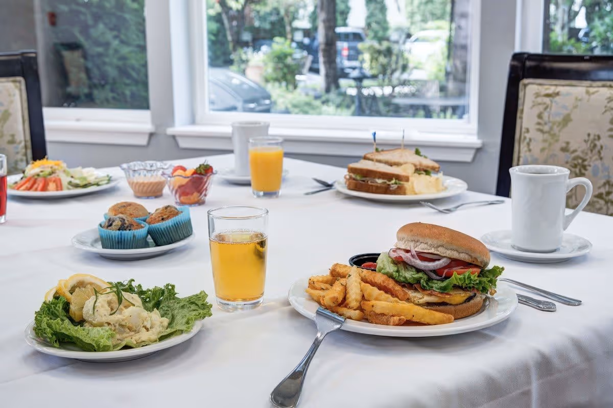 A dining table set with various plates of food including a sandwich with lettuce, tomato, and onion served with crinkle-cut fries, a plate of potato salad with lemon slices on lettuce, a plate with muffins, a sandwich with potato chips, a glass of orange juice, a glass of iced tea, and a white coffee cup. The table is covered with a white tablecloth and is near a window showing greenery outside.