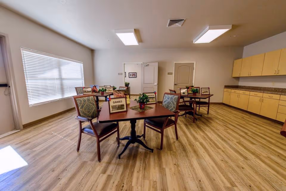 Bright communal activity room with several tables and chairs, potted plants on the tables, cabinets along the right wall and a large window on the left.