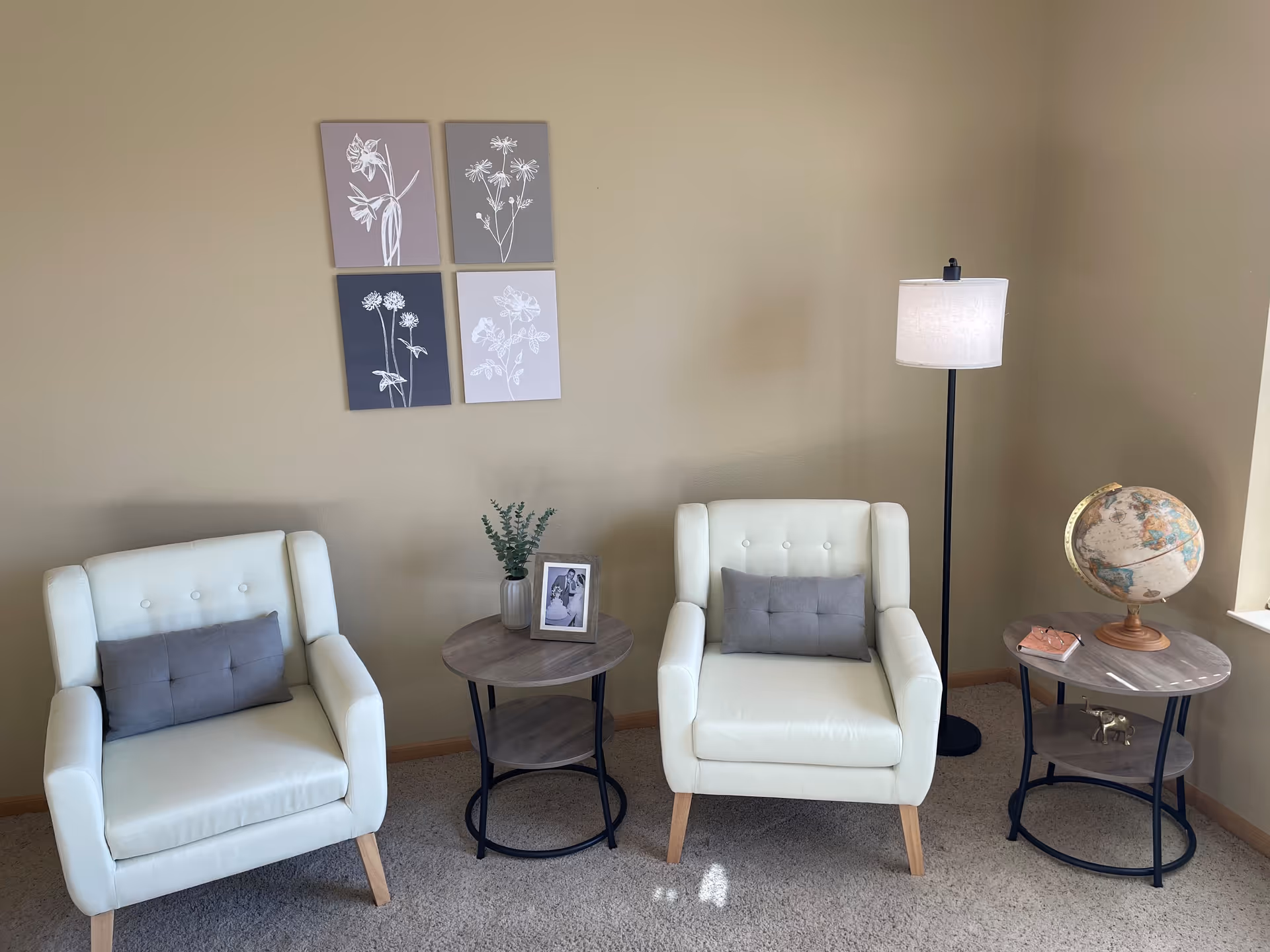 A cozy sitting area with two white cushioned armchairs, each with a gray pillow. Between the chairs is a small round wooden side table with a potted plant and a framed photo. To the right of the right chair is a floor lamp and another round wooden side table holding a globe and a small decorative item. Four botanical prints are hung on the beige wall behind the chairs.