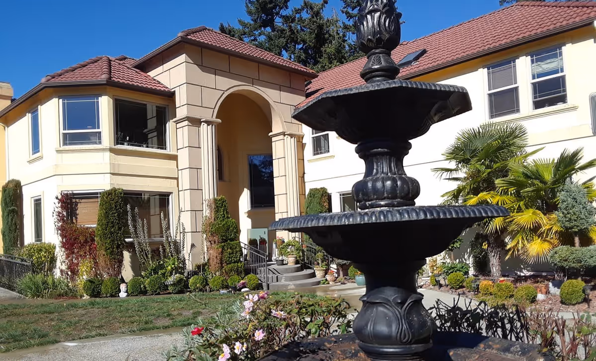 Exterior view of a two-story building with beige walls and red-tiled roof, featuring a large arched entrance. In the foreground, there is a black decorative fountain surrounded by a garden with various plants and flowers. The sky is clear and blue.