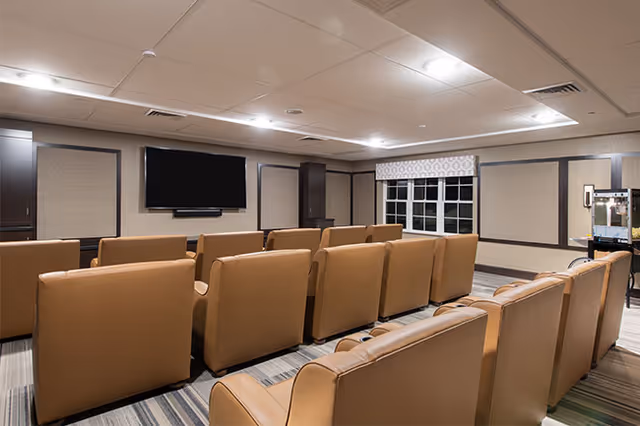 A cozy media room with multiple rows of tan leather chairs facing a large flat-screen TV mounted on the wall. The room has a striped carpet, beige walls with dark trim, a window with a patterned valance, and ceiling lights providing soft illumination.