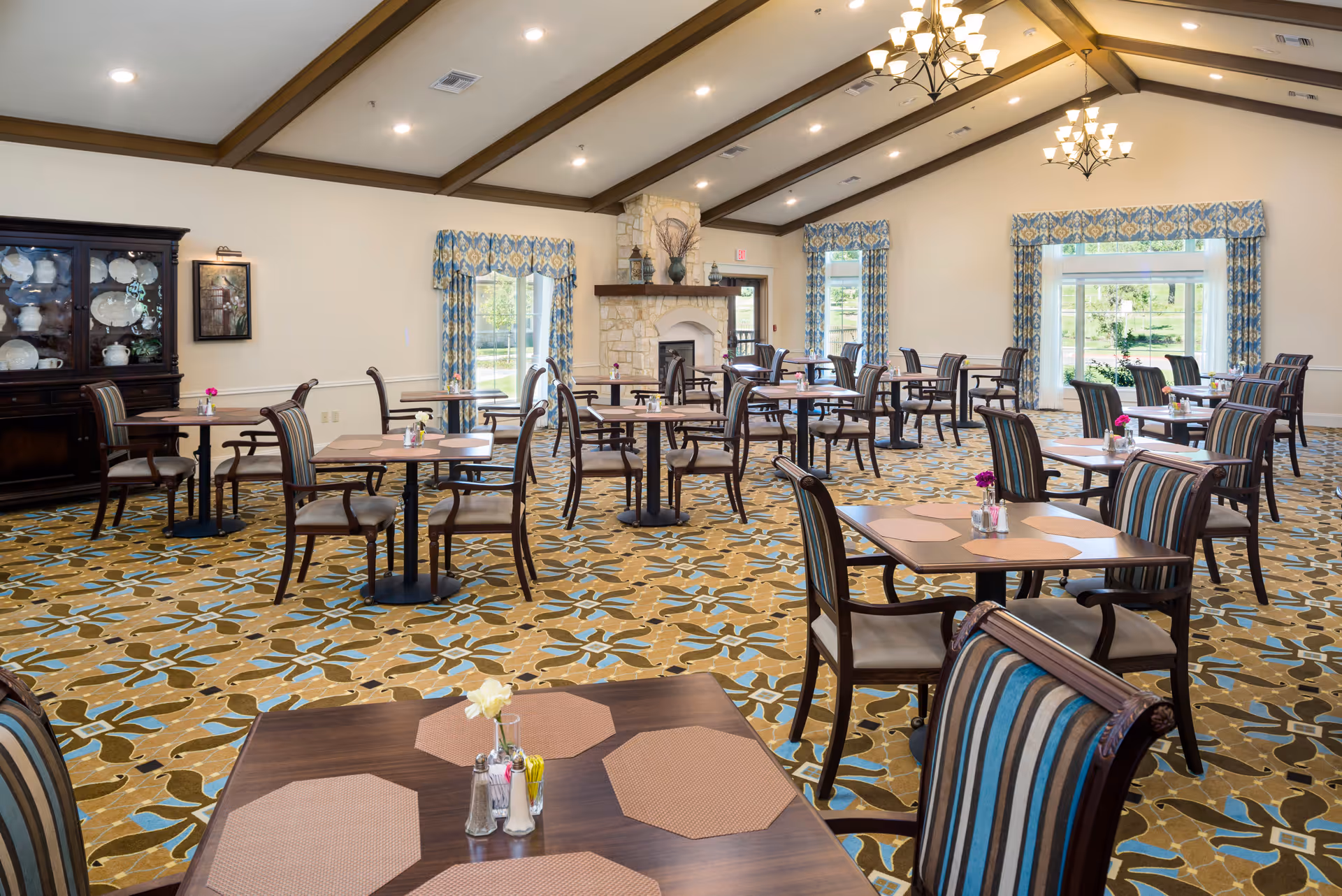 A spacious dining room with multiple wooden tables and striped cushioned chairs arranged neatly. The room features a patterned carpet in shades of brown, blue, and beige, large windows with blue and beige patterned curtains, and a stone fireplace at the far end. The ceiling has recessed lighting and two chandeliers.