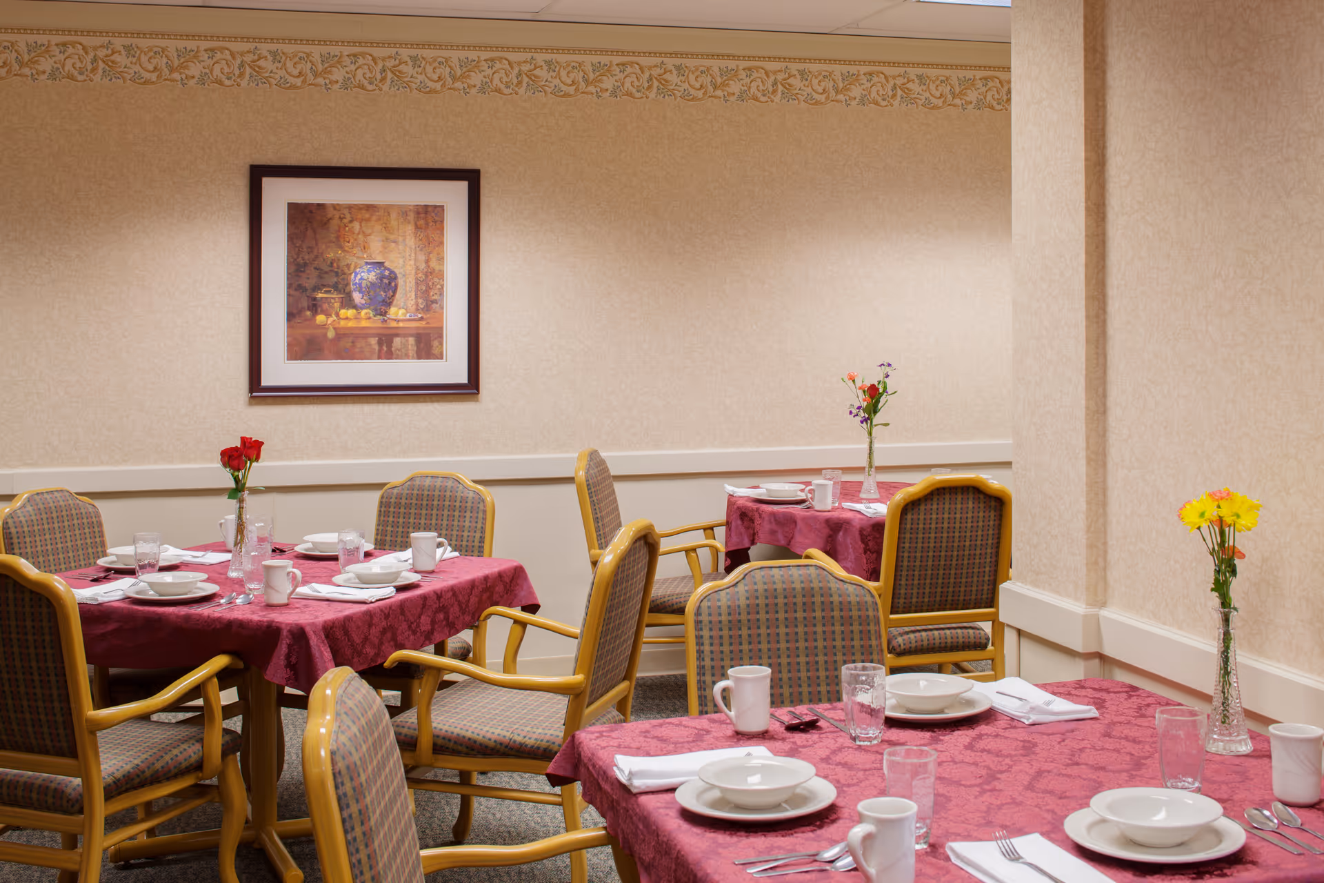 Dining room with tables covered in burgundy tablecloths, set with dishes and glassware, surrounded by upholstered chairs and small vases of flowers.