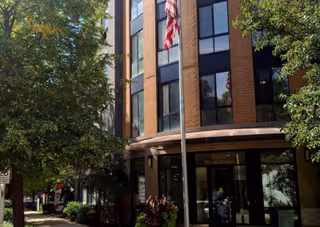 Front exterior of a brick senior living building with glass doors, an American flag, and surrounding trees.