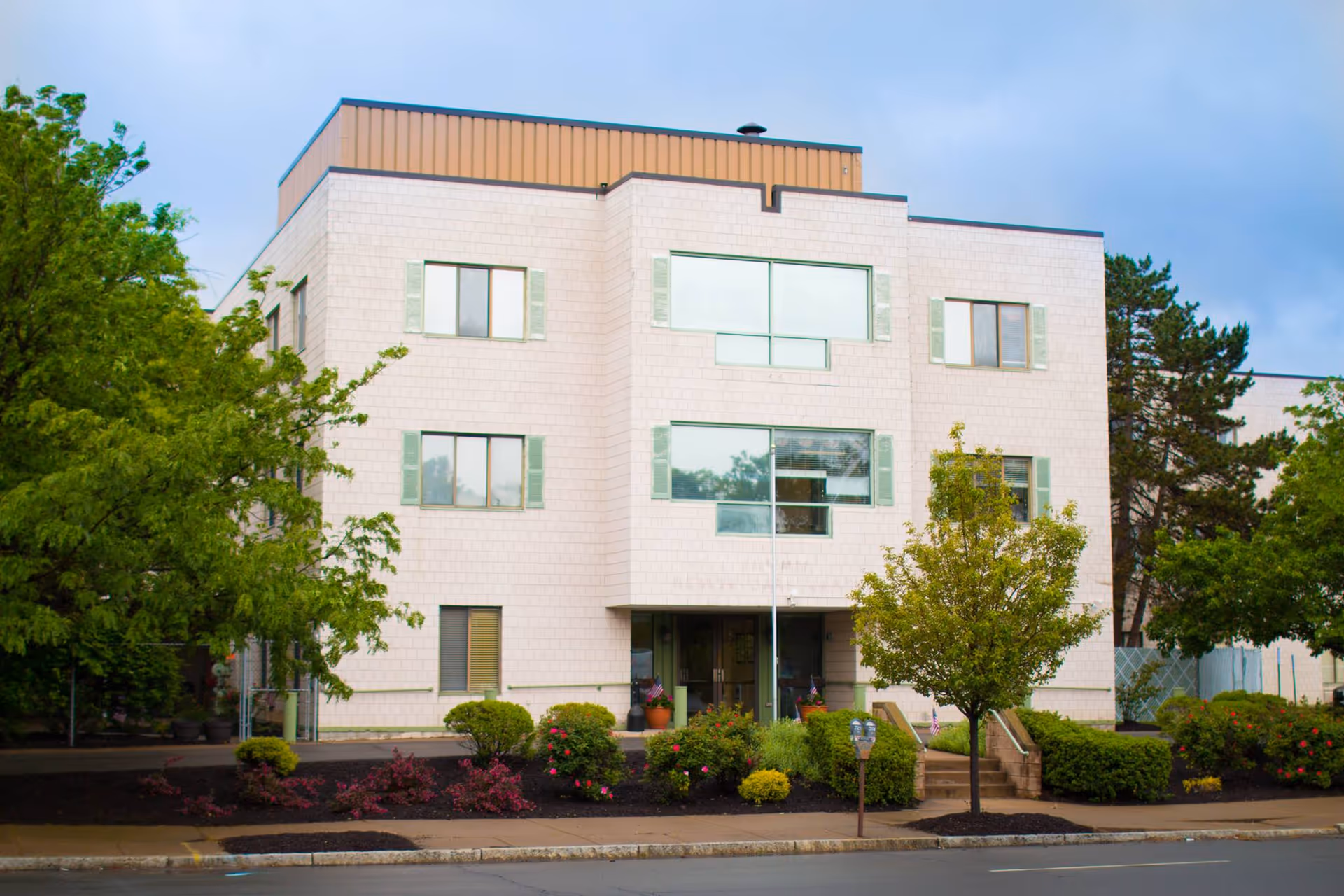 Exterior view of a three-story light-colored brick building with multiple windows and green shutters. There are trees and landscaped bushes in front of the building, along with a sidewalk and a street in the foreground. The sky is partly cloudy.
