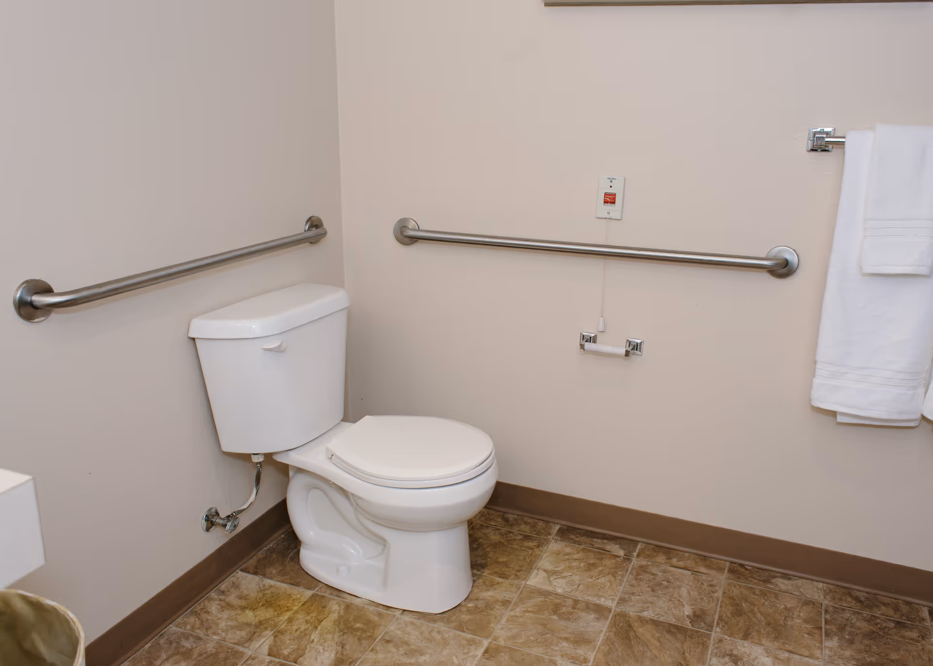 A clean bathroom with a white toilet, beige walls, and tiled floor. There are two stainless steel grab bars on the walls for support, a white towel hanging on a towel rack, and a pull cord emergency alarm above the toilet paper holder.