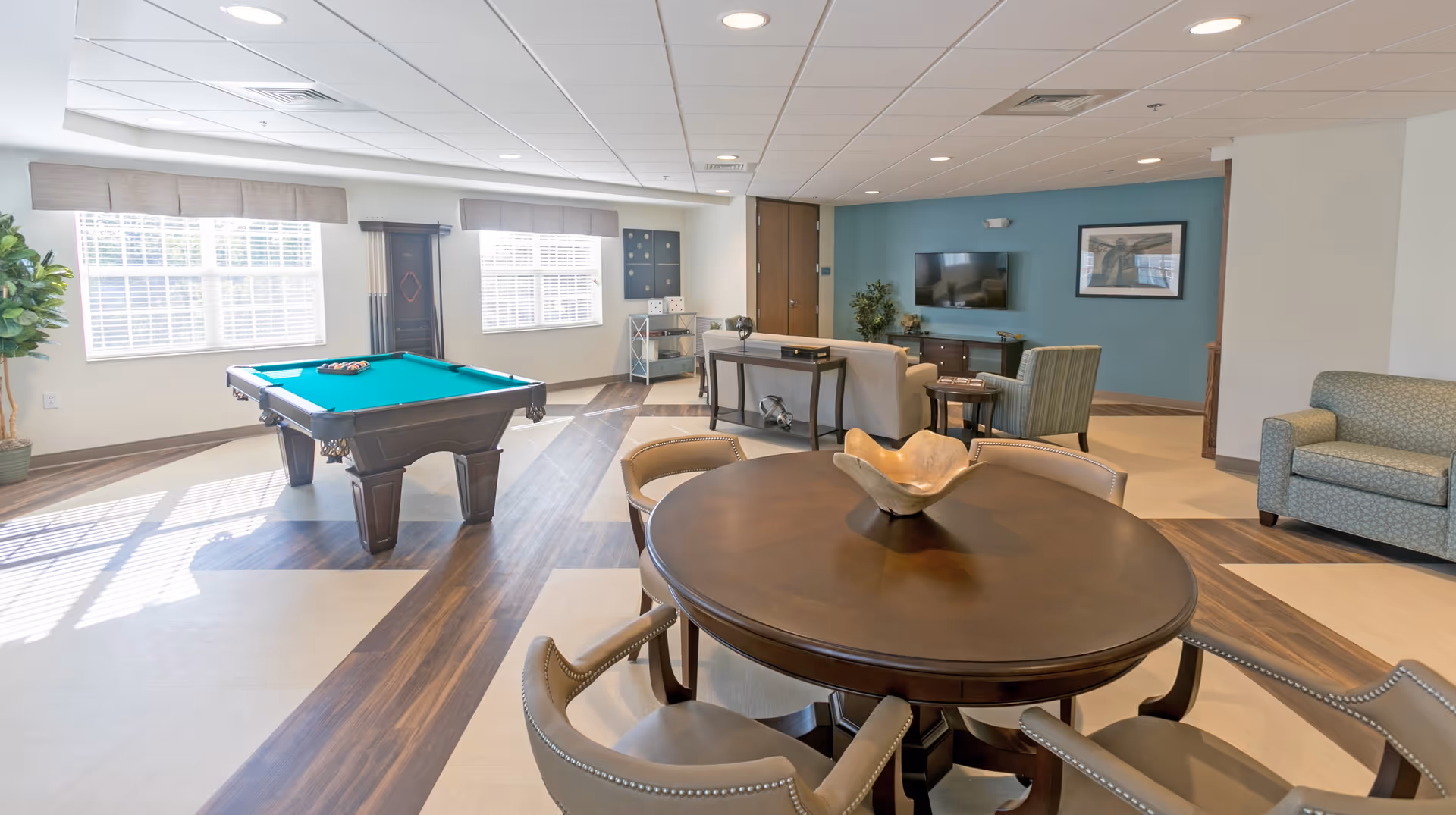 A bright and spacious common area in a senior living facility featuring a pool table near two large windows with blinds, a round wooden table with six upholstered chairs, a beige sofa, an armchair, a wall-mounted TV on a blue accent wall, and decorative plants and artwork.