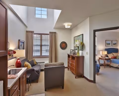 Interior view of a senior living facility suite with a small kitchenette on the left, a seating area with a couch and armchair in the center, and a bedroom visible through an open doorway on the right. The room has high ceilings with a large window, beige walls, and carpeted floors. There is a wooden dresser with decorative items and a wall clock on the far wall.