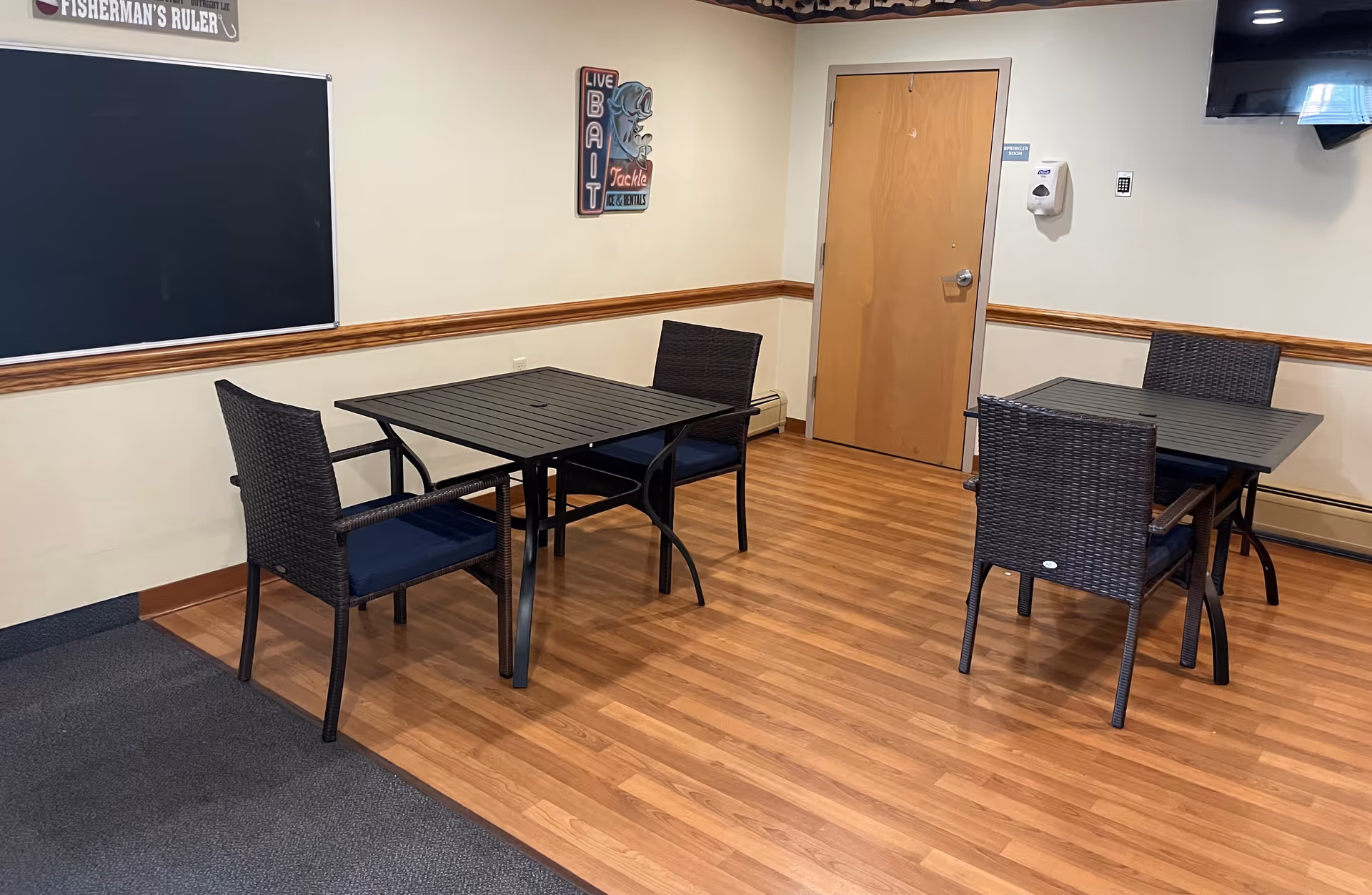 A small room with two black square tables and four dark wicker chairs with blue cushions. The room has light-colored walls with a wooden chair rail, a wooden door, a blackboard on one wall, and a wall-mounted television. The floor is a combination of wood and carpet.