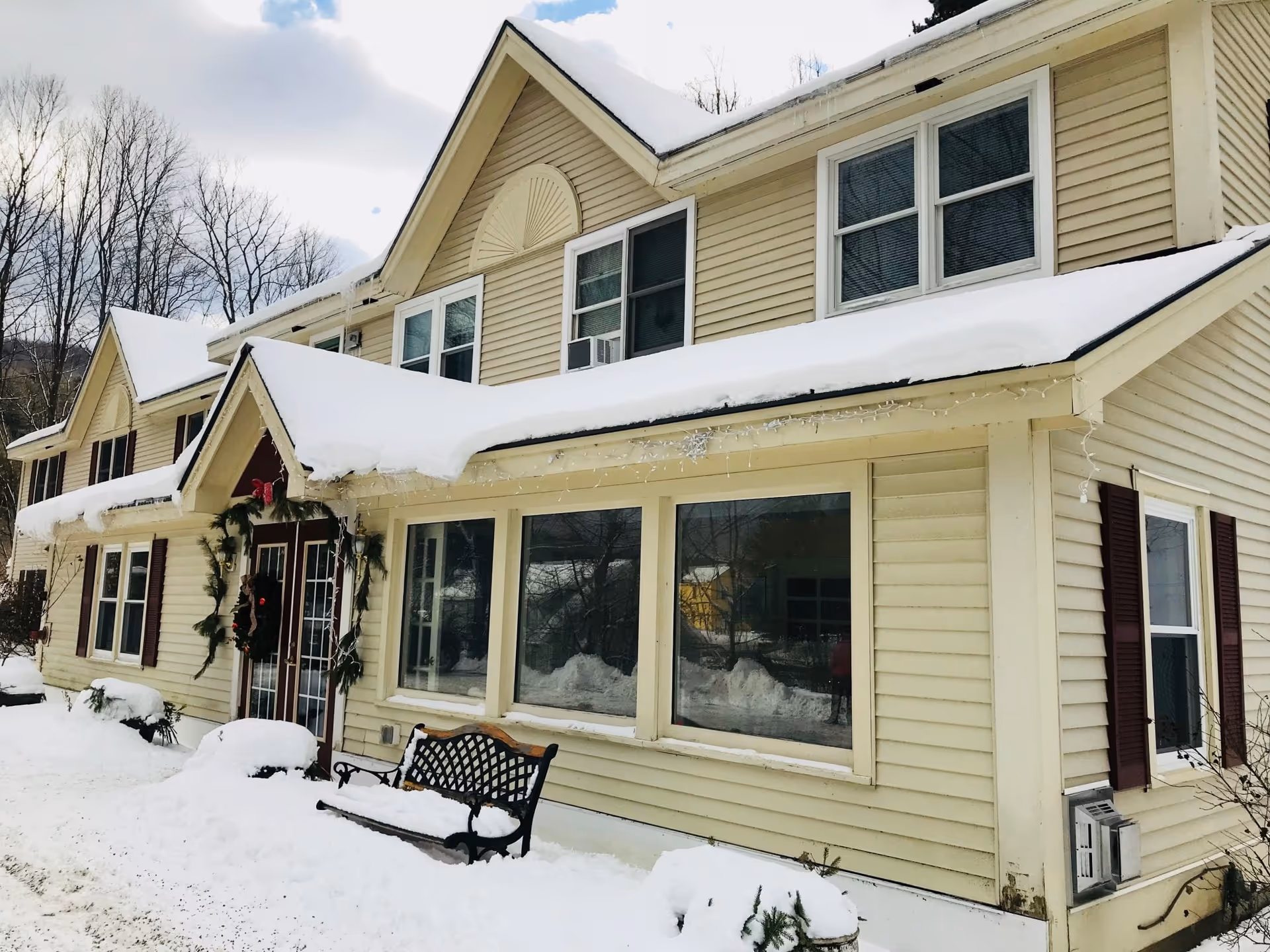 Exterior view of a beige two-story building with snow-covered roof and ground. The building has multiple windows, a bench covered in snow near the entrance, and holiday decorations around the door. Leafless trees are visible in the background under a cloudy sky.
