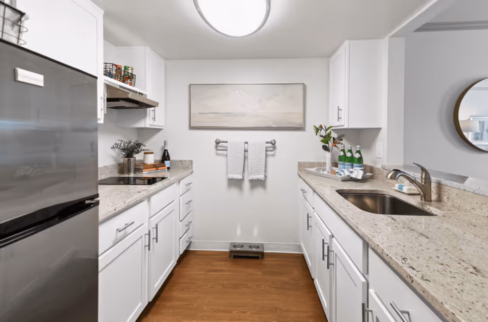 A modern kitchen with white cabinets and granite countertops. On the left side, there is a stainless steel refrigerator and a stovetop with a range hood above it. On the right side, there is a sink with a faucet and a tray holding bottles and a vase with flowers. A towel rack with two white towels is mounted on the wall at the center, below a framed landscape painting. The floor is wooden, and a pet feeding station is placed beneath the towel rack.