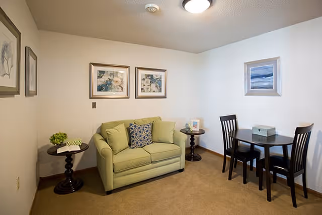 A small living area with a light green loveseat adorned with three cushions, two side tables with decorative items, and a round wooden dining table with two chairs. The walls are decorated with framed artwork, and the room has beige carpeting and a white ceiling with a light fixture.