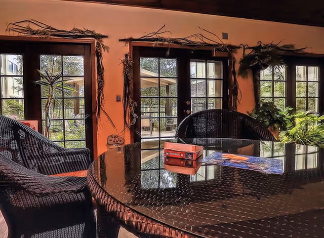 Sunlit common room with wicker chairs and a round glass-top table in front of French doors opening to a patio.