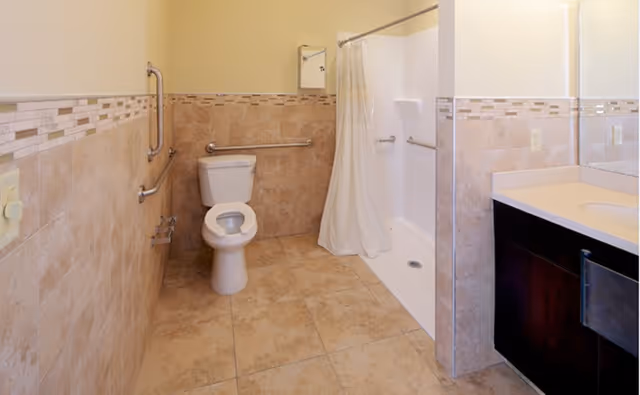 Tiled accessible bathroom featuring a toilet with grab bars, a roll-in shower with curtain, and a vanity sink.