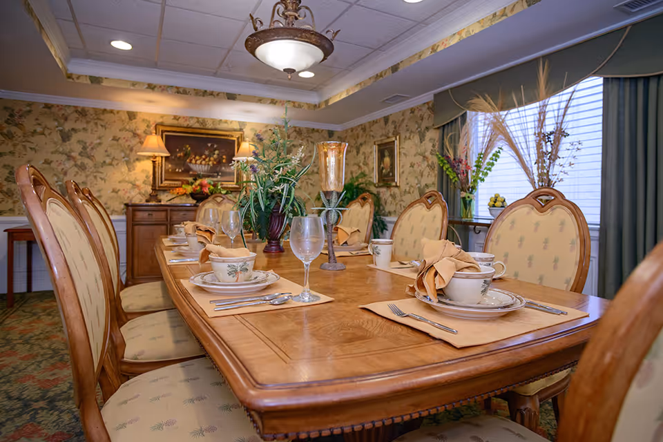 A formal dining room with a long wooden table set for a meal. The table has place settings with plates, cups, napkins, and glasses. The chairs have cushioned seats and backs with a patterned fabric. The room features floral wallpaper, a sideboard with lamps and a painting, and a window with green curtains and decorative plants on the windowsill.