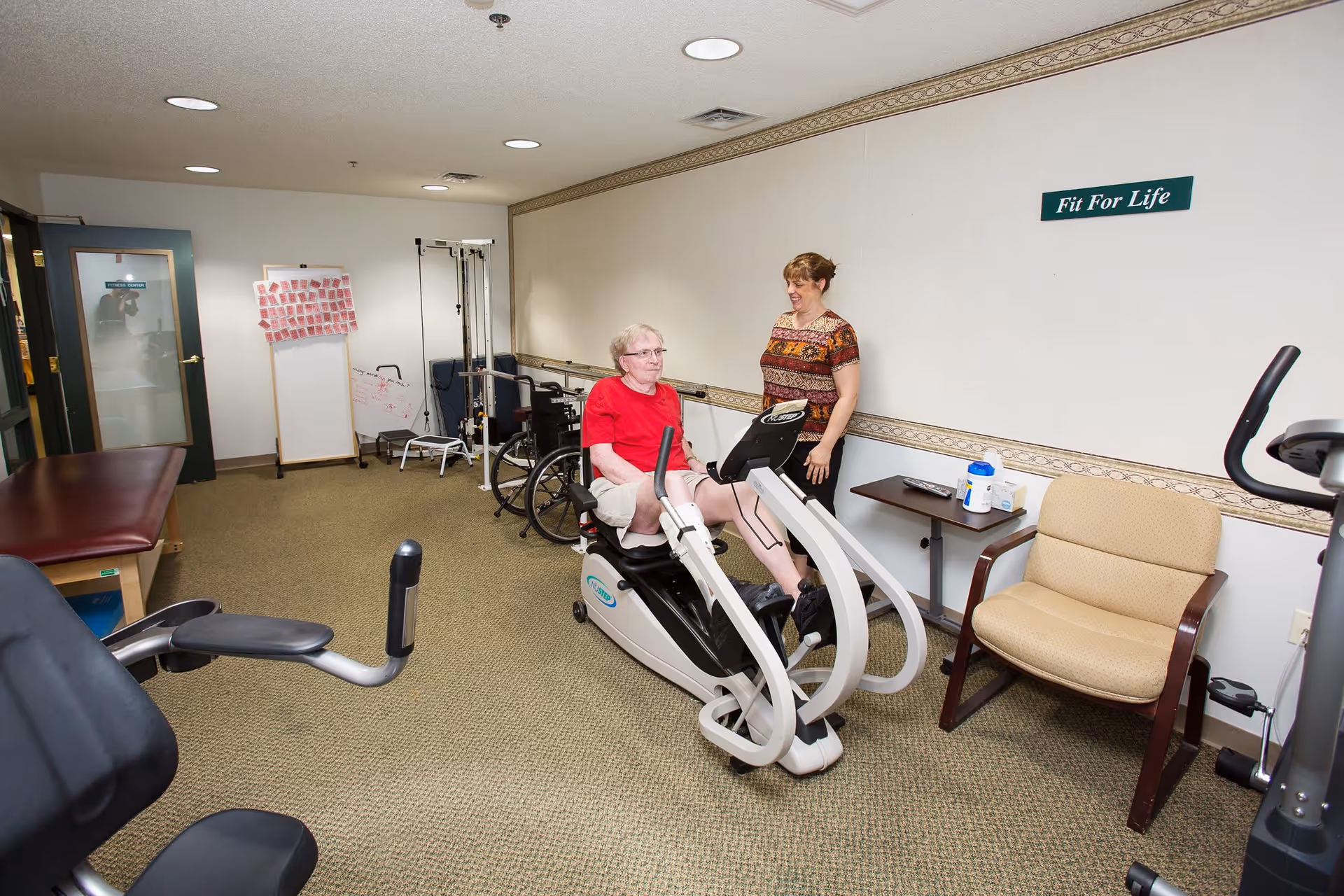 An elderly man in a red shirt exercises on a recumbent bike in a fitness room while a woman stands beside him smiling. The room has exercise equipment, a wheelchair, chairs, and a sign on the wall that reads 'Fit For Life'.