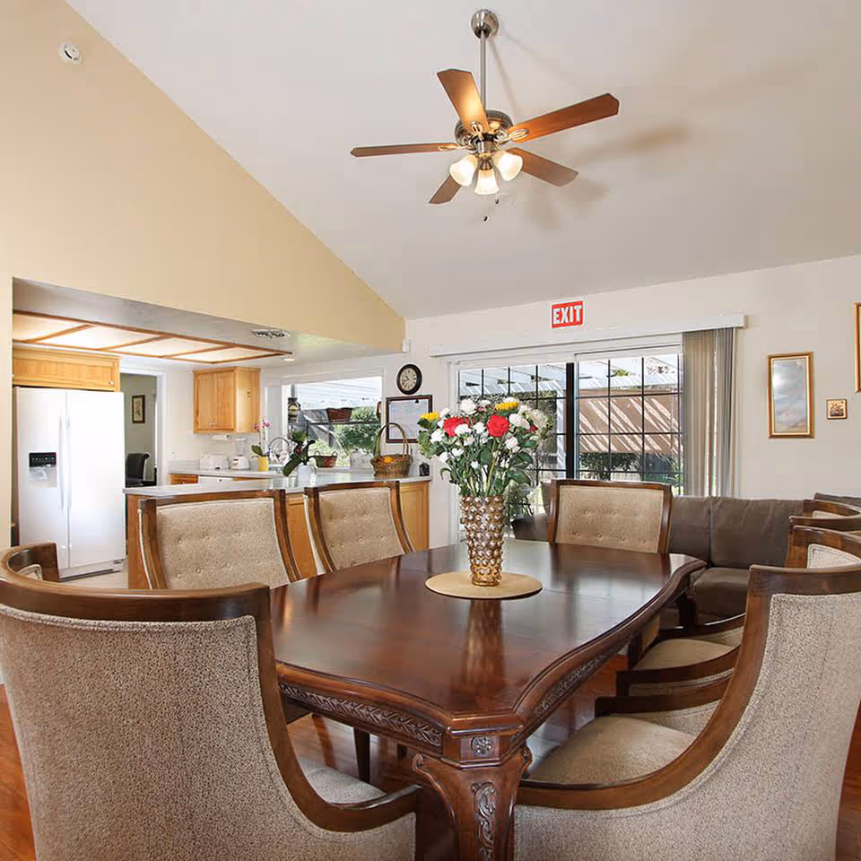 A dining area with a polished wooden table surrounded by cushioned chairs. A vase with colorful flowers is placed in the center of the table. In the background, there is a kitchen with wooden cabinets, a white refrigerator, and a ceiling fan with lights above the dining table. Large sliding glass doors with vertical blinds lead to an outdoor area, and an exit sign is visible above the doors.