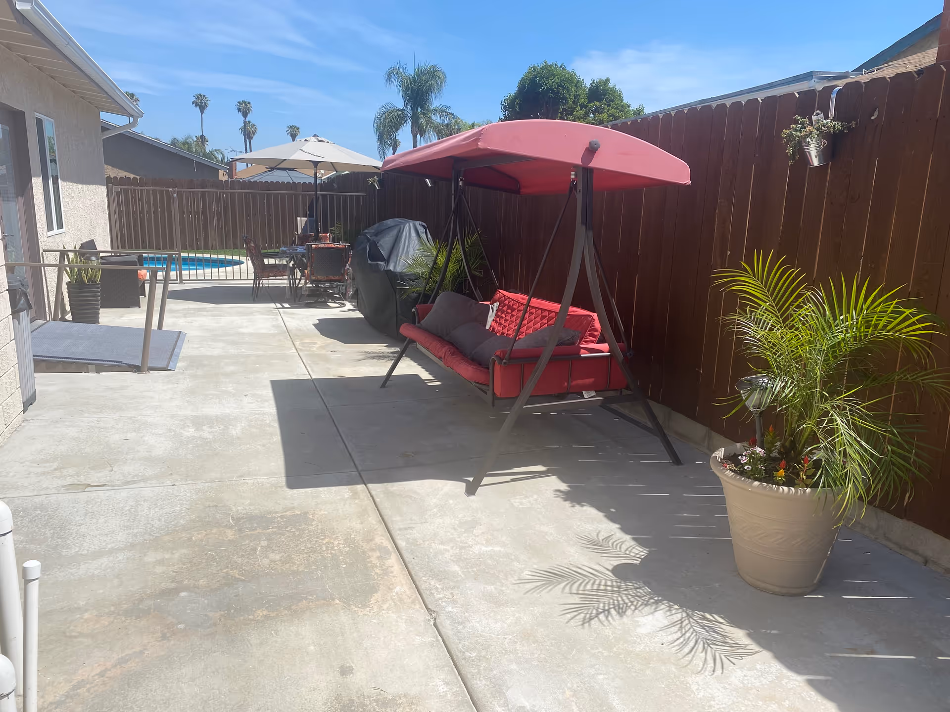 Outdoor patio area with a red cushioned swing with a canopy, a potted plant, a barbecue grill covered with a black cover, a table with chairs under a large umbrella, and a swimming pool in the background. The area is enclosed by a wooden fence and there are palm trees visible beyond the fence under a clear blue sky.
