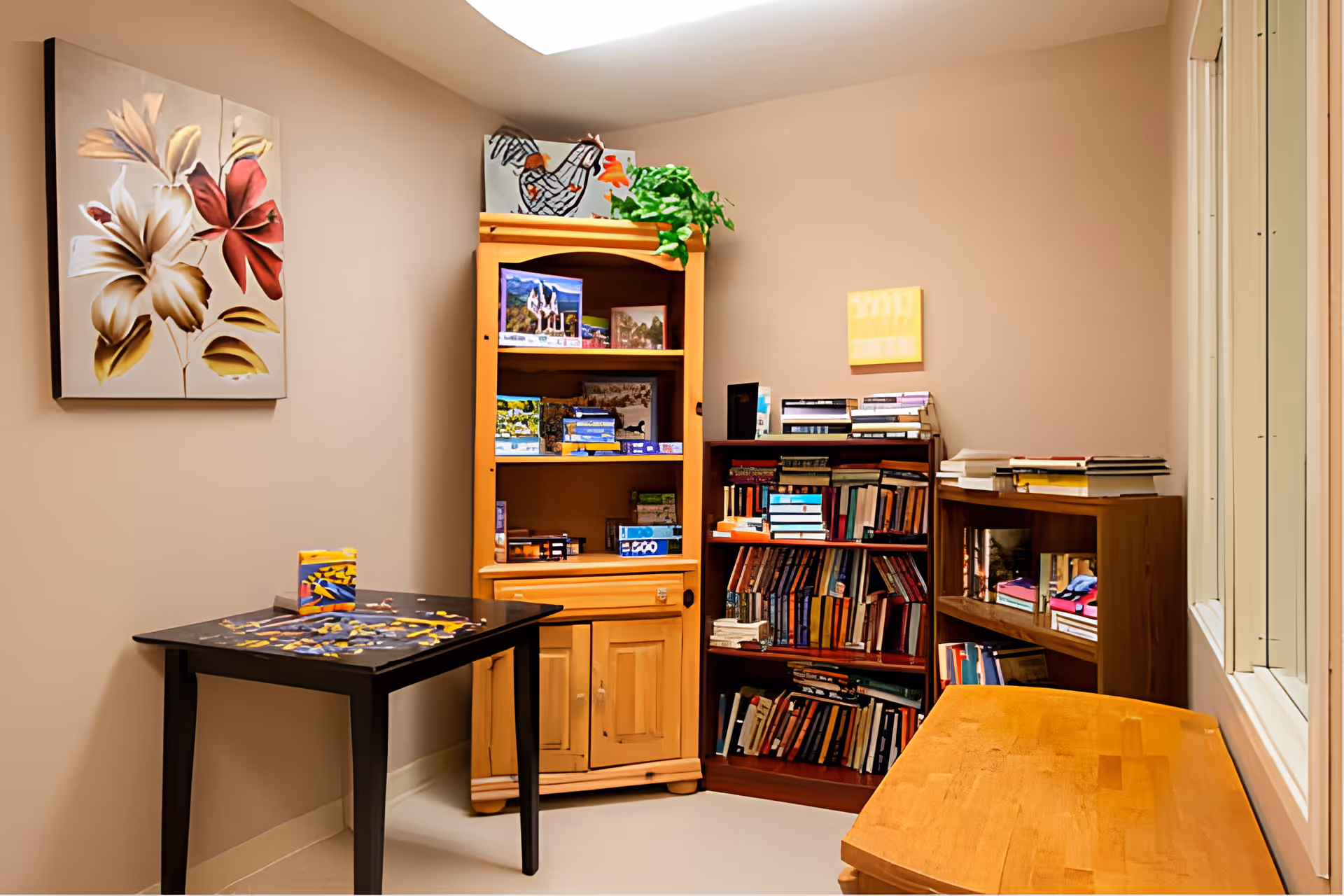 A cozy room with beige walls featuring a black table with a puzzle in progress, three wooden bookshelves filled with books and games, a small green plant on top of one shelf, and a floral painting on the wall.